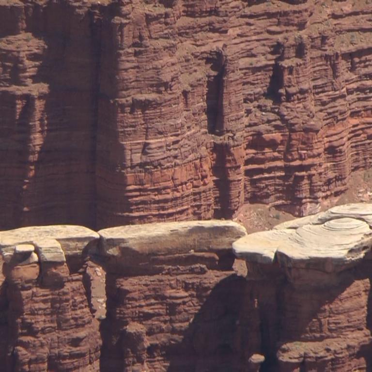 Massive, gestufte Sandsteinwände eines Canyons mit erodierten Felsblöcken im Vordergrund.