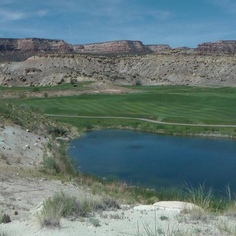 Eine weite Landschaft mit einem Teich im Vordergrund, einem Golfplatz in der Mitte und roten Felsformationen im Hintergrund.