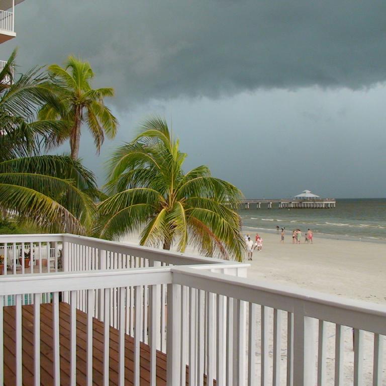 Blick auf einen tropischen Strand von einem Balkon aus, eingerahmt von Palmen und unter einem dramatischen, bewölkten Himmel.