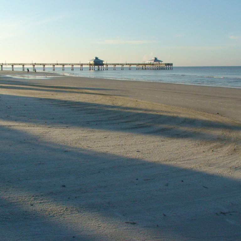 Ein langer Holzpier erstreckt sich vom Sand über das ruhige Meer hinweg, während die Sonne lange Schatten auf den Strand wirft.