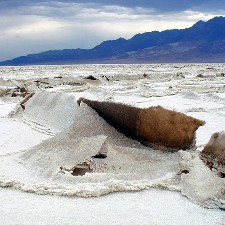 Weite, flache Salzebene mit weißen Ablagerungen im Vordergrund und einer Bergkette am Horizont unter bewölktem Himmel.