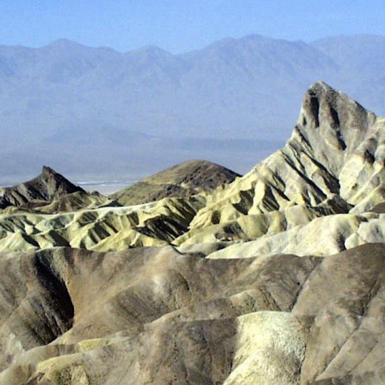 Ausgedehnte Wüsten- oder Hochlandlandschaft mit sanften Hügeln und zerklüfteten Felsformationen vor einer Bergkette.