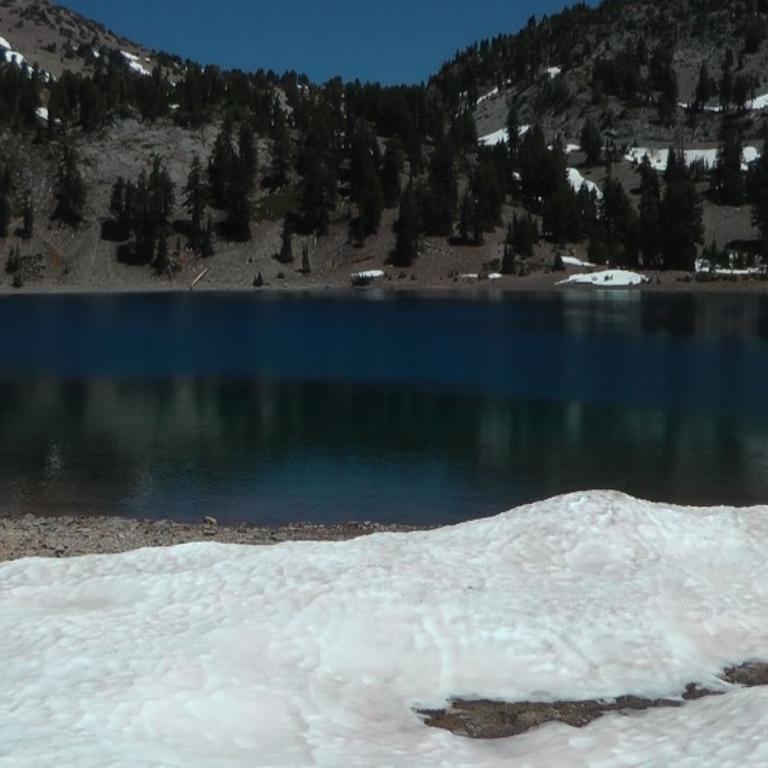 Ein ruhiger Bergsee mit bewaldetem Ufer und verschneitem Vordergrund.
