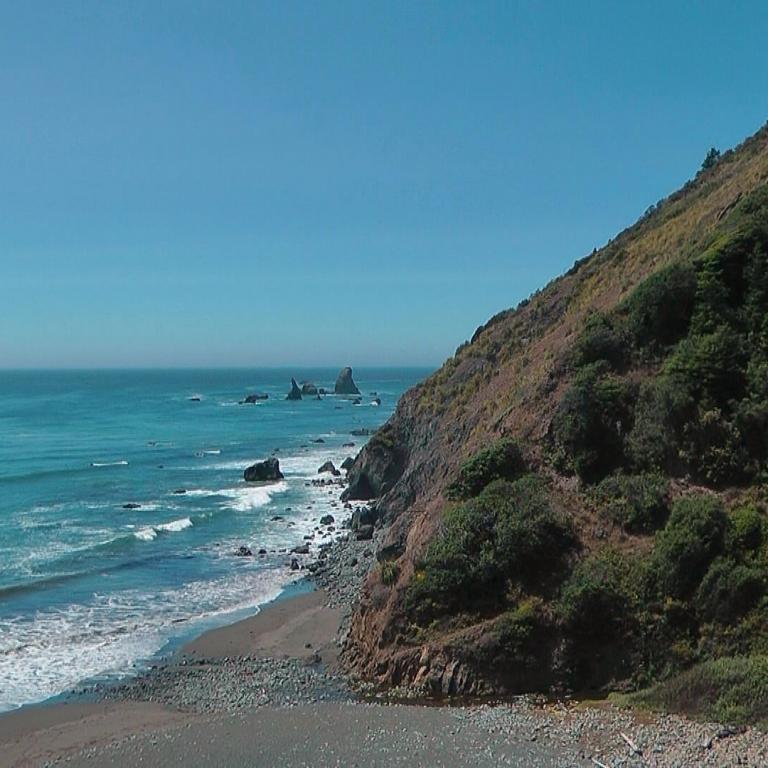 Steile, bewaldete Klippe, die zu einem schmalen Strand und zum türkisfarbenen Meer hinabfällt.