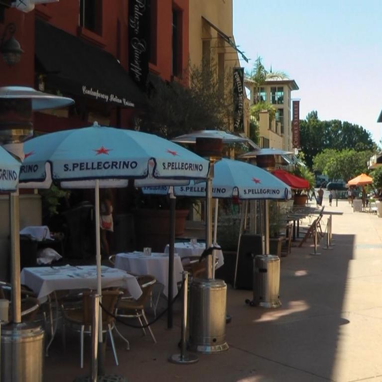 Eine belebte Straßenszene mit Cafés, die mit blauen und grünen Markisen und weiß gedeckten Tischen ausgestattet sind.