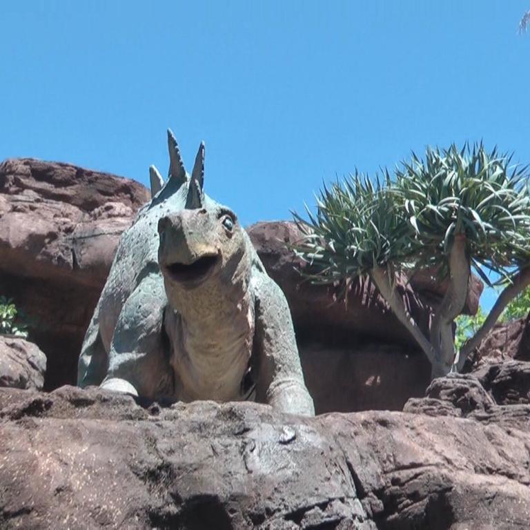 Eine große Skulptur eines Dinosauriers liegt auf dunklen Felsen unter einem klaren blauen Himmel.