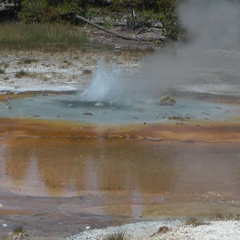 Ein Wasserbecken mit aufsteigendem Dampf inmitten einer natürlichen, spärlich bewachsenen Landschaft.