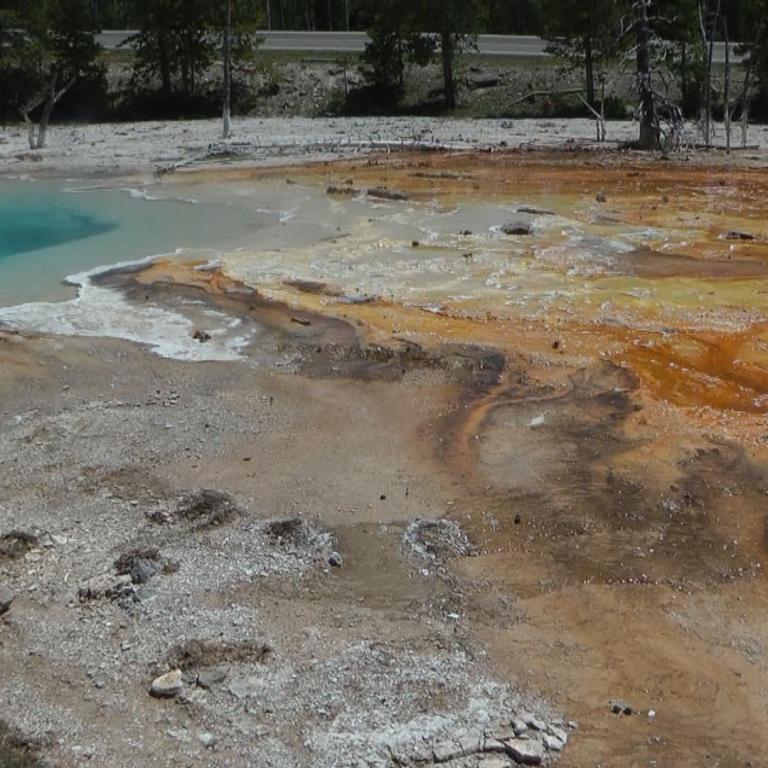 Ein Geothermalgebiet mit einem türkisfarbenen Wasserbecken und rotbraunen, mineralhaltigen Ablagerungen am Ufer.