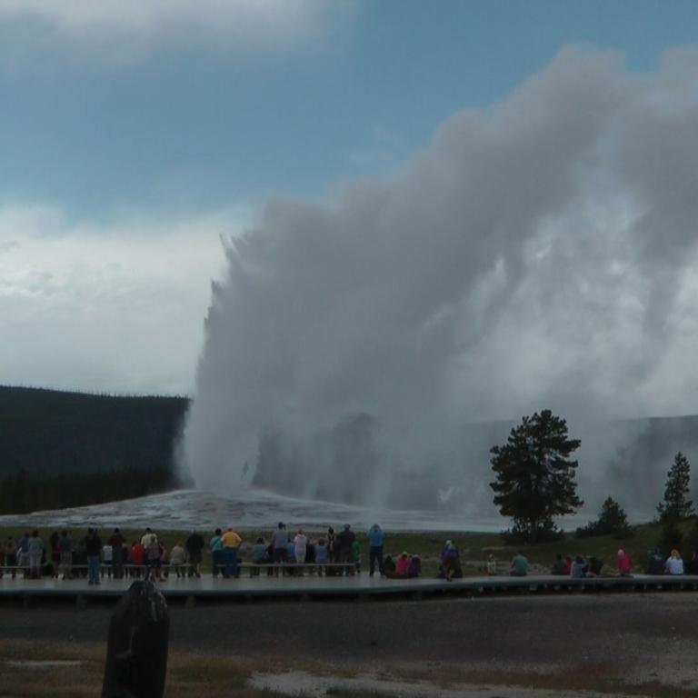 Ein massiver Wasserstrahl steigt aus einer geothermischen Quelle auf und erzeugt eine große Dampfwolke. Menschen beobachten das Spektakel von einem Au