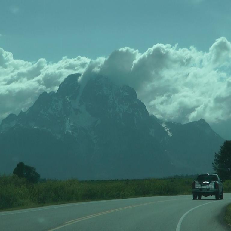 Ein Auto fährt auf einer asphaltierten Straße vor einer Kulisse majestätischer, wolkenverhangener Berge.