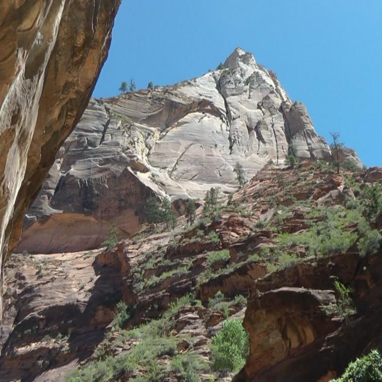 Ein dramatischer Ausblick auf einen tiefen Canyon, begrenzt von hohen Felswänden und dominiert von einem steilen, hellen Bergpeak unter blauem Himmel.