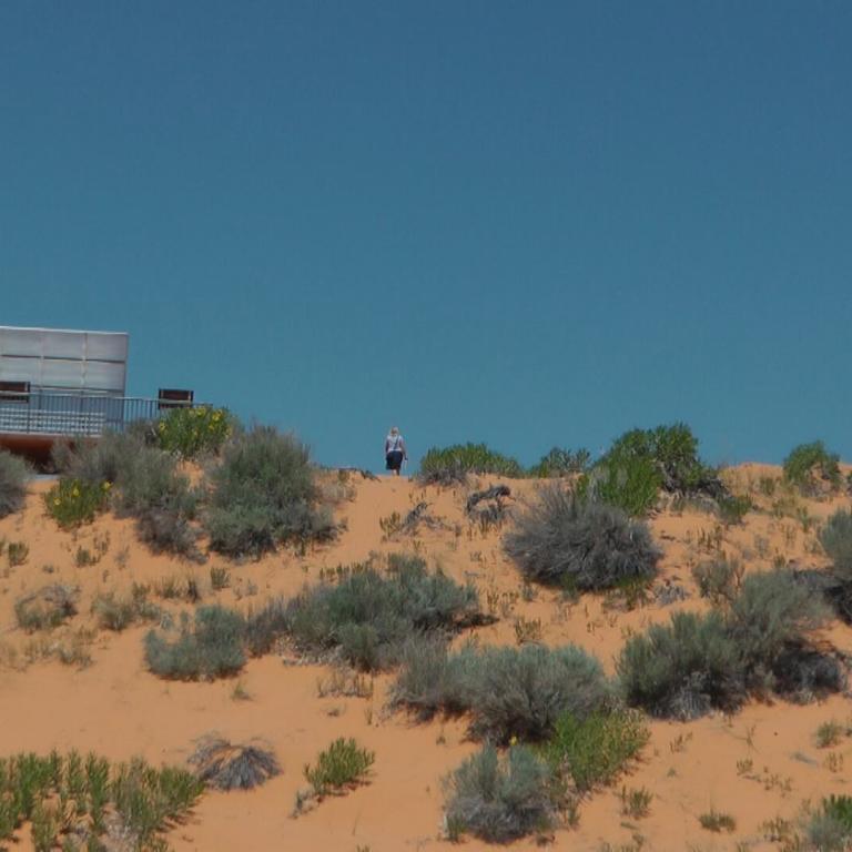 Eine moderne, erhöhte Plattform inmitten einer kargen Dünenlandschaft unter blauem Himmel.