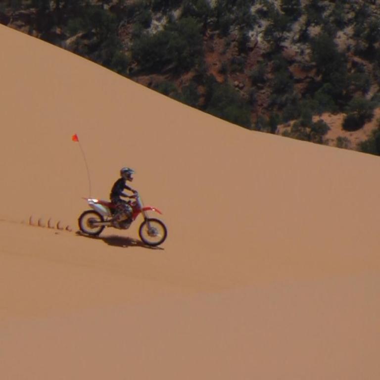 Ein Motorradfahrer fährt eine große, sandige Dünenfläche hinunter, im Hintergrund sind bewaldete Hügel zu sehen.