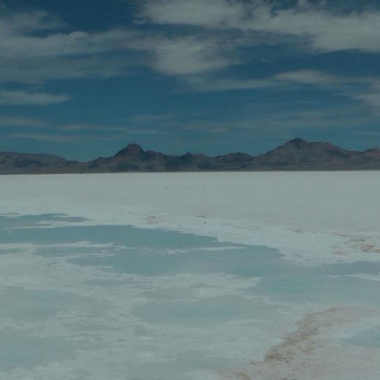 Eine weite, flache Ebene, die wie ein Salzsee wirkt, mit Bergketten am Horizont und einem bewölkten Himmel.