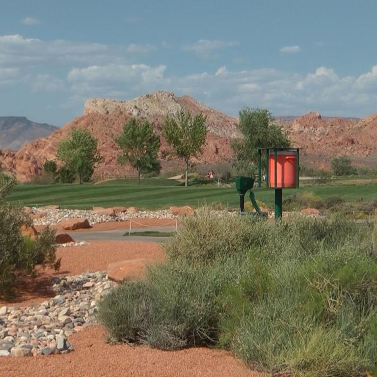 Panoramaaufnahme eines Golfplatzes in einer ariden, bergigen Wüstenlandschaft mit Buschwerk im Vordergrund.