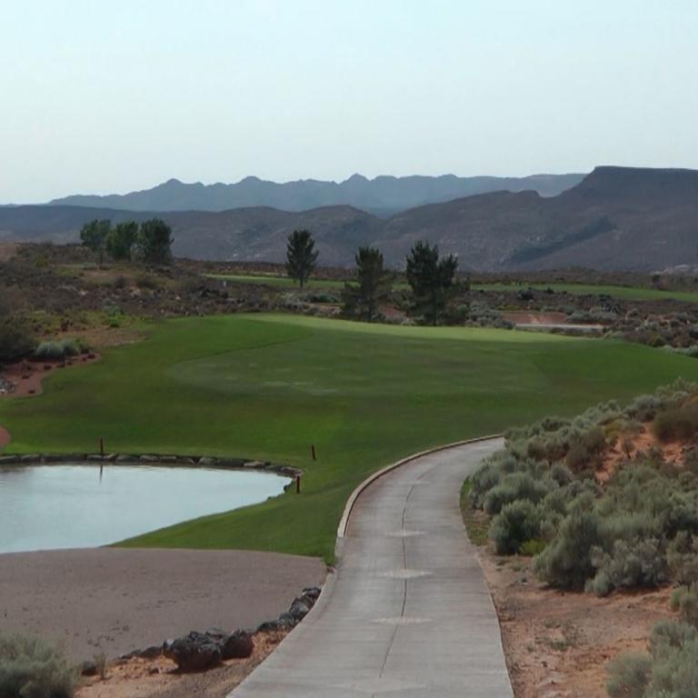 Panoramablick auf einen Golfplatz im Tal mit einem Wasserbecken und Bergen im Hintergrund.