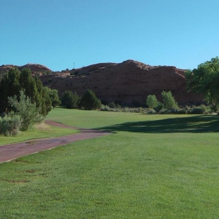 Ein Golfplatz mit einem Weg im Vordergrund und einem felsigen, hügeligen Hintergrund unter blauem Himmel.